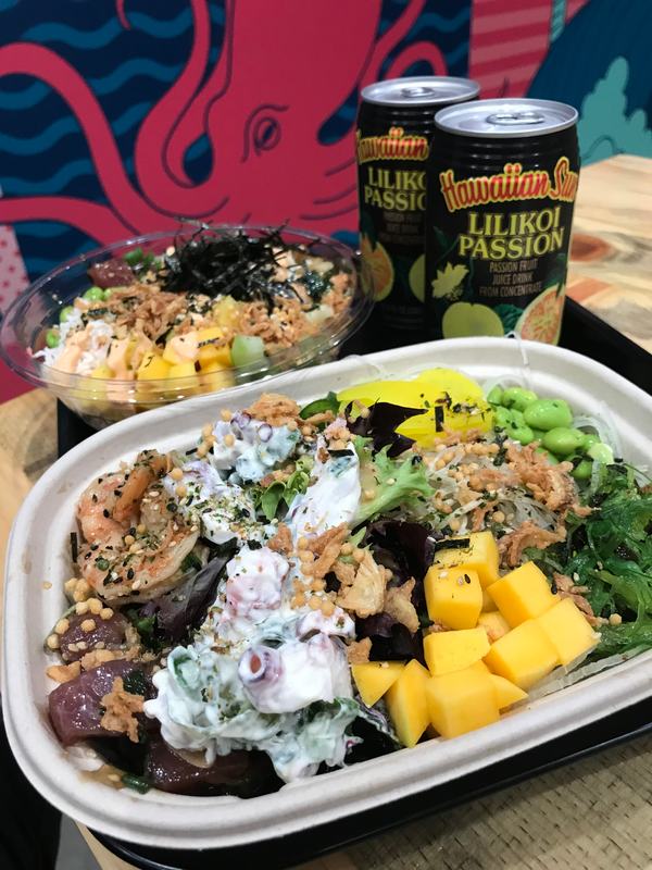 Poke bowl in a takeout tray with mango and edamame, cans of Lilikoi Passion and the octopus mural behind